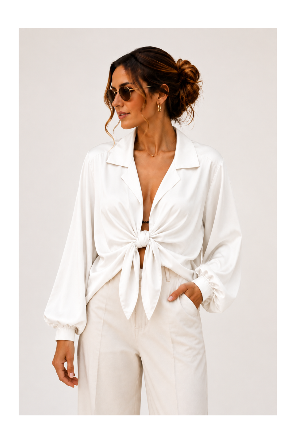 A woman wearing a white long-sleeve collared shirt tied at the front and white trousers, photographed against a plain light background.