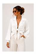 A woman wearing a white long-sleeve collared shirt tied at the front and white trousers, photographed against a plain light background.
