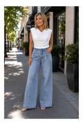 A woman stands on a city sidewalk wearing a white sleeveless cowl-neck top and light-wash wide-leg denim jeans, with her hand in a pocket. Storefronts, trees, and planters are visible in the background.