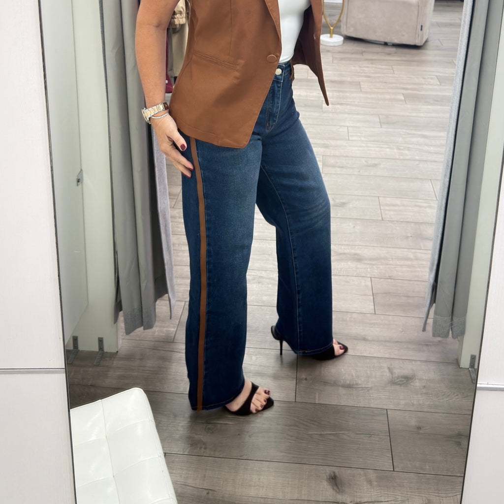 A woman takes a mirror selfie in a boutique wearing a sleeveless brown vest, a white tube top, dark blue wide-leg jeans with a brown side stripe, and black heeled sandals.