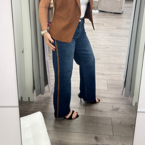 A woman takes a mirror selfie in a boutique wearing a sleeveless brown vest, a white tube top, dark blue wide-leg jeans with a brown side stripe, and black heeled sandals.