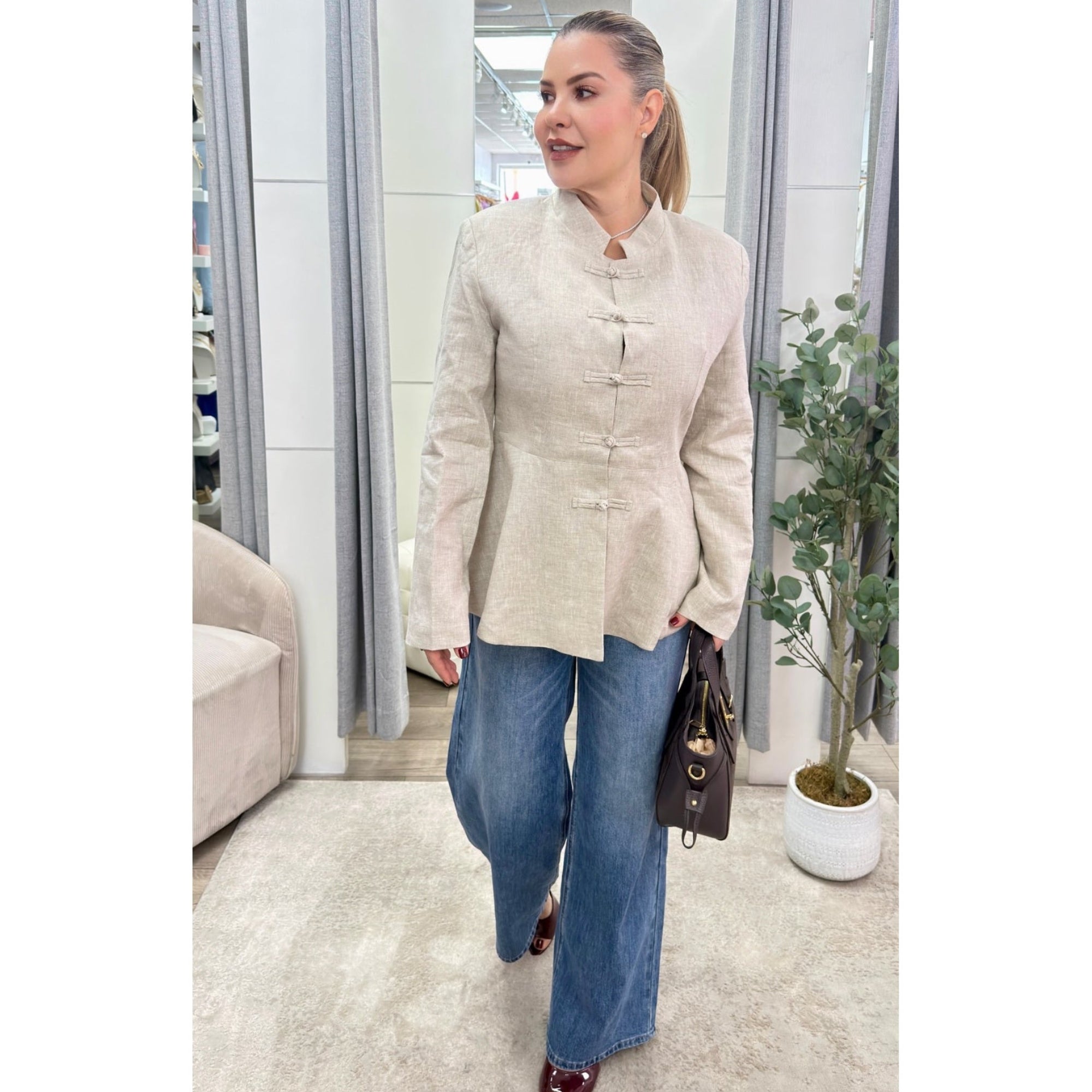 A woman modeling a beige textured long-sleeve jacket with traditional Chinese-style frog closures, paired with wide-leg blue jeans and holding a dark brown handbag.