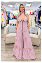 A woman modeling a pink and white vertically striped sleeveless jumpsuit with a large front bow and tiered wide legs, standing in a clothing boutique.