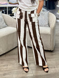 A woman posing in a clothing boutique, wearing a white graphic t-shirt with a brown and mint green mermaid design and high-waisted brown and white striped wide-leg pants.