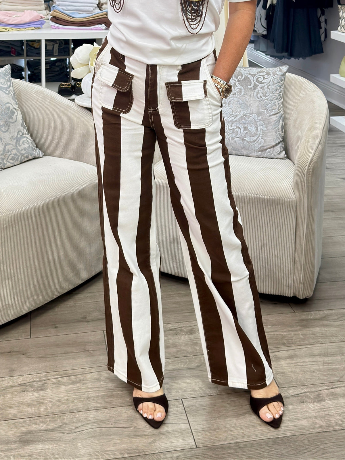 A woman posing in a clothing boutique, wearing a white graphic t-shirt with a brown and mint green mermaid design and high-waisted brown and white striped wide-leg pants.