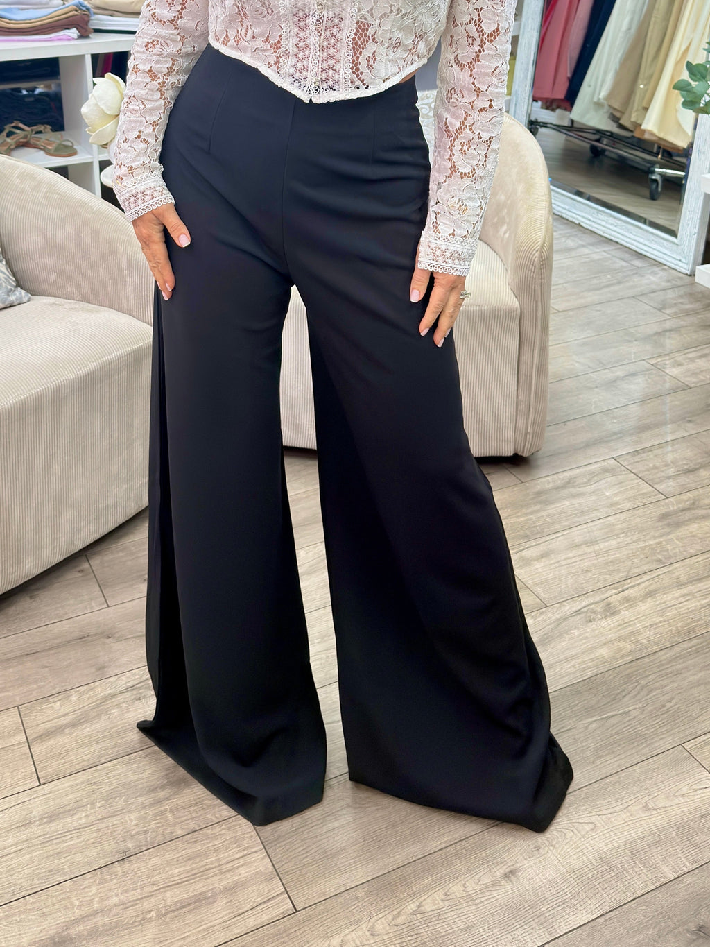 A woman modeling a white lace long-sleeve blouse paired with black wide-leg pants inside a clothing boutique.