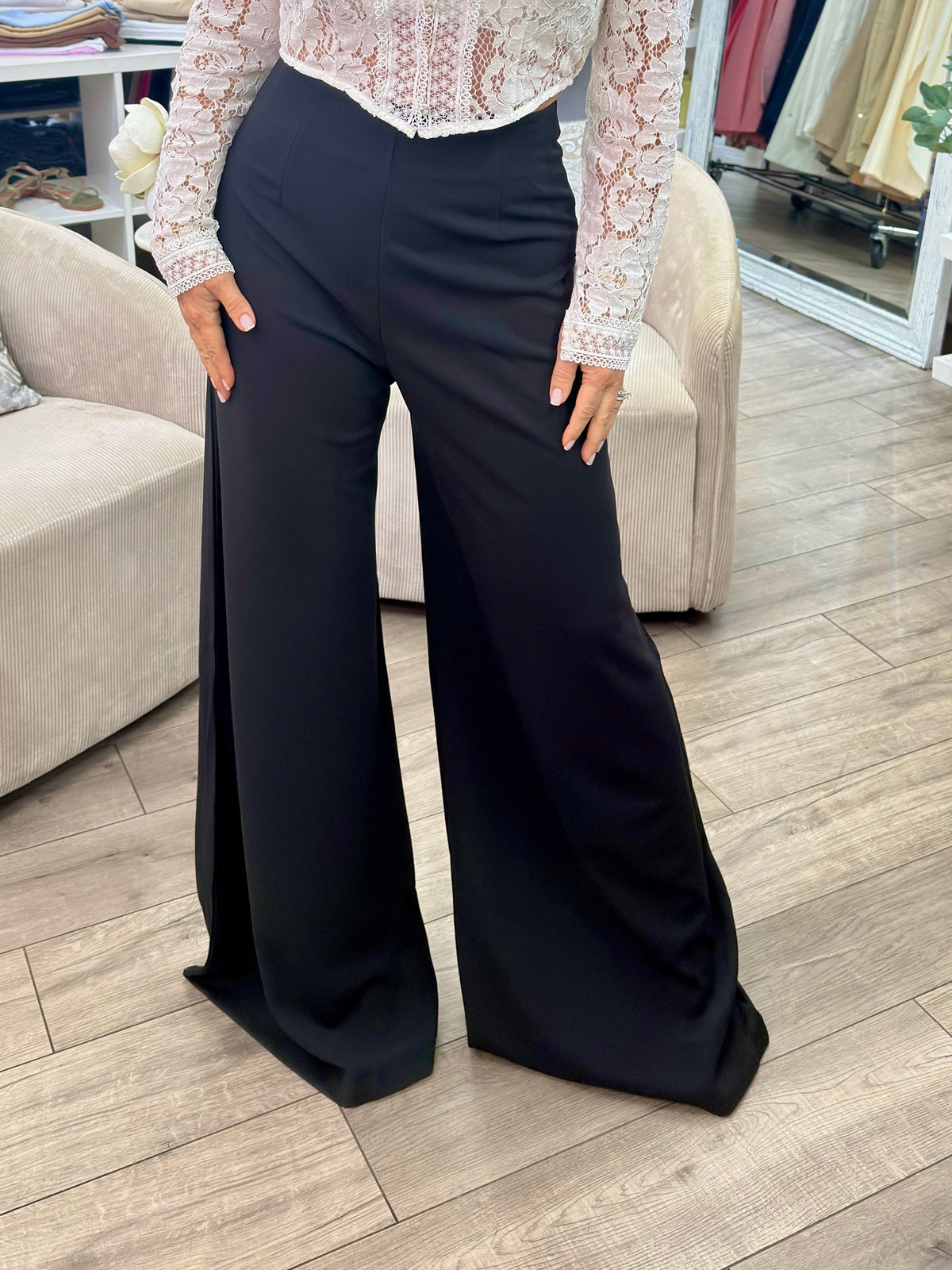 A woman modeling a white lace long-sleeve blouse paired with black wide-leg pants inside a clothing boutique.