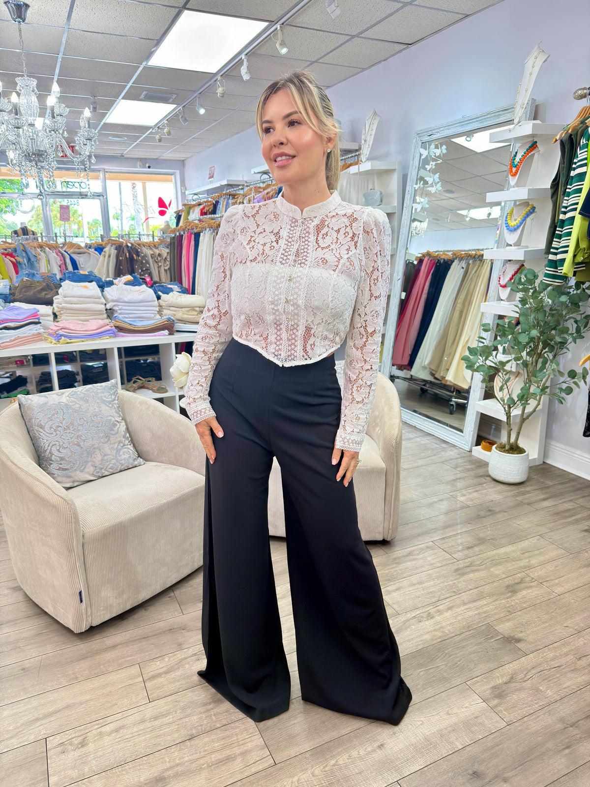 A woman modeling a white lace long-sleeve blouse paired with black wide-leg pants inside a clothing boutique.