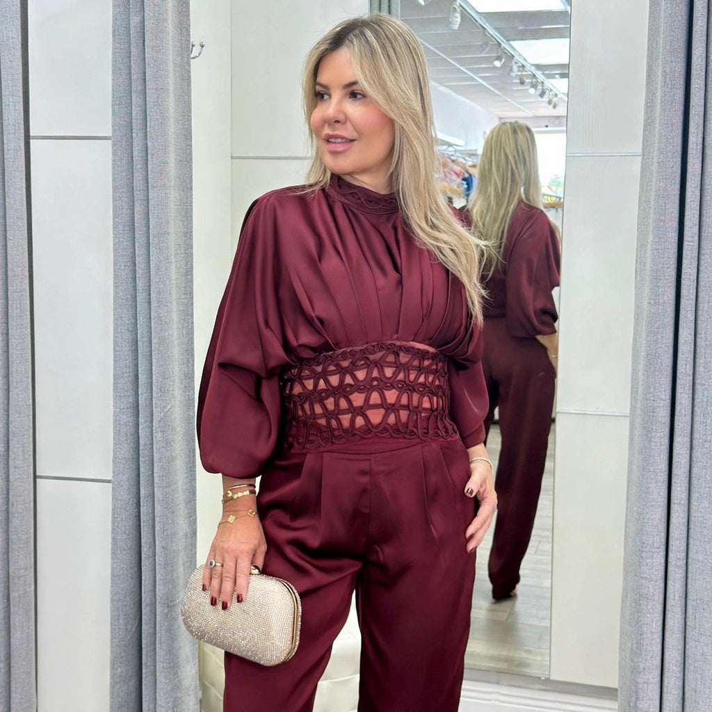 A woman modeling a burgundy satin jumpsuit with a high neck, balloon sleeves, and a lace-up waist, standing in a fitting room with gray curtains and a mirror.