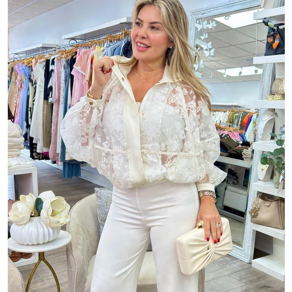 A woman models a white lace blouse with puffed sleeves and white wide-leg trousers in a boutique setting, holding a matching cream clutch.