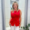 A woman modeling a sleeveless bright red button-front top paired with white wide-leg pants, standing in a modern boutique fitting room with gray curtains and a potted plant.