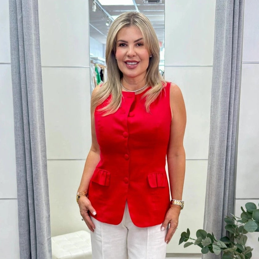 A woman modeling a sleeveless bright red button-front top paired with white wide-leg pants, standing in a modern boutique fitting room with gray curtains and a potted plant.