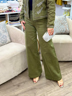 A woman models an olive green two-piece set consisting of a cropped jacket with silver embroidery and matching wide-leg pants inside a boutique clothing store.