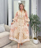 A woman modeling a long, flowing kaftan-style dress with floral prints and ornate gold embroidery, standing in a modern indoor setting with a mirror, curtains, and a potted plant.